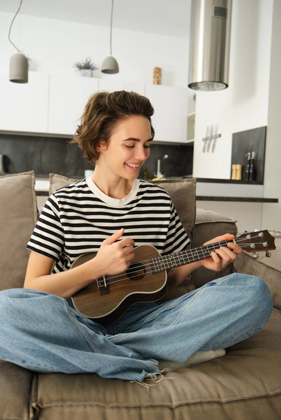 Vertical portrait of happy young woman playing ukulele, learns new musical instrument, sits on sofa at home, smiling with joy. - Photo, Image