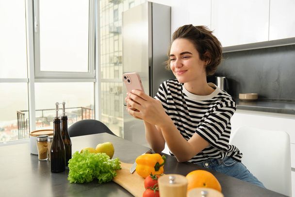 Tatlı gülümseyen bir kadının portresi salata pişiriyor, akıllı telefona bakıyor, yemek tarifi izliyor, yemek hazırlıyor, sebze doğruyor, mutfakta yemek yapıyor. Sağlıklı beslenme ve yaşam tarzı kavramı - Fotoğraf, Görsel