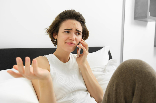 Portret van een vrouw die een telefoongesprek voert met een vriend, luistert naar een oproepbare persoon en haalt zijn schouders op, hoort ingewikkelde situaties, ligt in bed en praat. - Foto, afbeelding