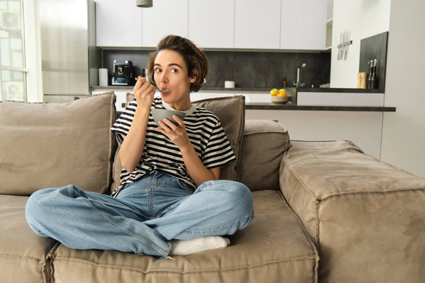 Портрет стильной молодой женщины на диване, кушающей злаки, держащей миску и ложку, смотрящей телевизор и завтракающей, проводящей время дома в гостиной. - Фото, изображение