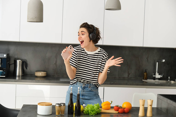 Retrato de la mujer cantando y bailando mientras cocina comida en la cocina, sosteniendo el teléfono inteligente, picando verduras en el mostrador de la cocina. - Foto, imagen