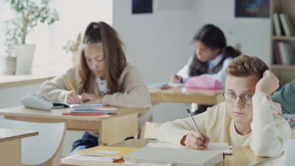 Concentration sélective de nerd fatigué garçon assis au bureau dans la salle de classe prendre des notes puis regarder la caméra pendant la leçon - Séquence, vidéo