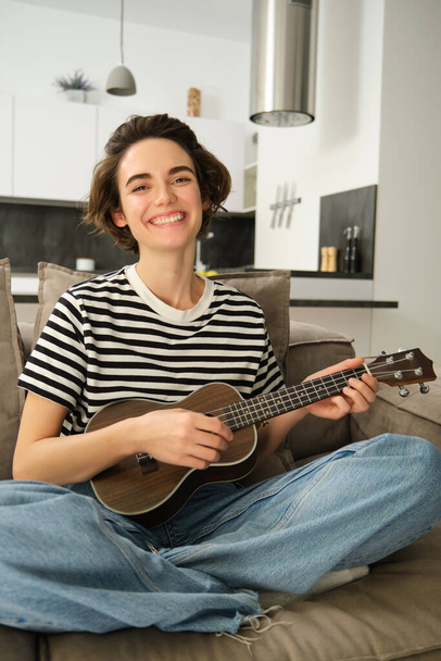 Vertical shot of young musician, woman learns how to play ukulele, sits on sofa with crossed legs and smiling at camera. - Photo, Image