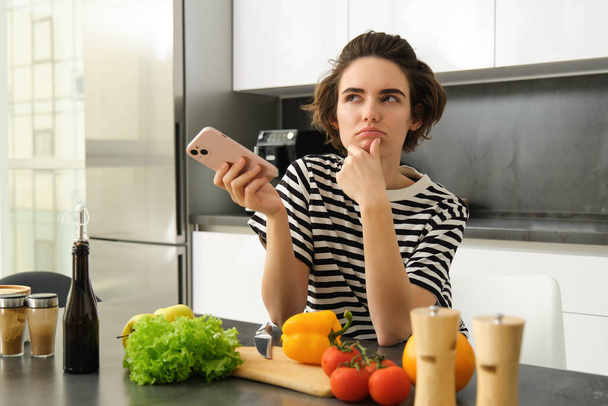 Genç bir kadının portresi mutfakta elinde sebzeler ve doğrama tahtasıyla duruyor, elinde akıllı telefon, cep telefonu tarayıcısında sağlıklı beslenme tarifleri arıyor, evde yemek pişiriyor.. - Fotoğraf, Görsel