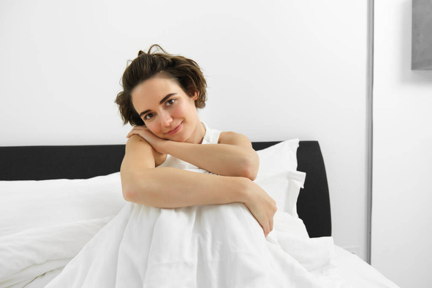 Image of beautiful brunette woman sitting on bed, covered in white linen sheets, looking at camera with dreamy, sensual smile, gazing at someone. Morning and lifestyle concept - Photo, Image
