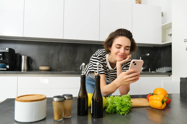 Menina cozinhar refeição saudável na cozinha, ler receita no smartphone, usando telefone celular e fazer refeição de legumes verdes frescos. - Foto, Imagem