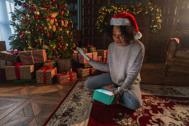 Joyeux Noël. Femme afro-américaine déballer ouvrir boîte cadeau près de l'arbre de Noël. Fille dans le salon avec arbre de Noël et cheminée boîte cadeau d'ouverture avec visage surprise. Veille de Noël à la maison - Photo, image