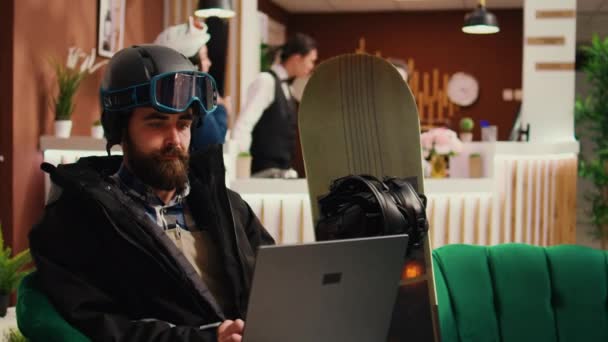Young adult checks website on laptop before going down on slopes, travelling for skiing leisure activity and winter sport. Hotel guest wearing protective helmet and professional gear. - Footage, Video