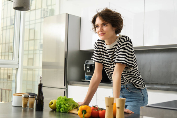 Porträt einer schönen jungen Frau, weibliches Model posiert am Schneidebrett mit Gemüse und Salatdressing, macht sich leichte Mahlzeit, veganes Essen zum Frühstück, steht in der Küche. - Foto, Bild