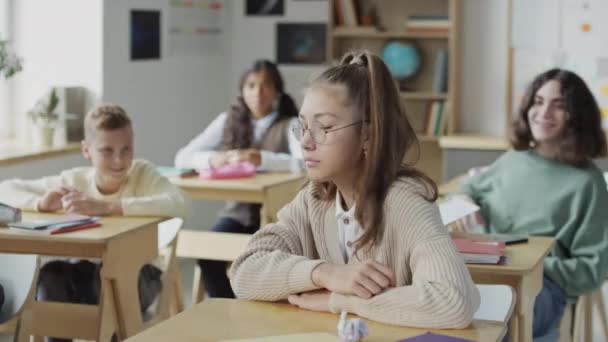 Selectieve focus shot van etnisch diverse middelbare scholieren zitten aan bureaus in klaslokaal pesten depressief meisje, het gooien van papieren naar haar - Video