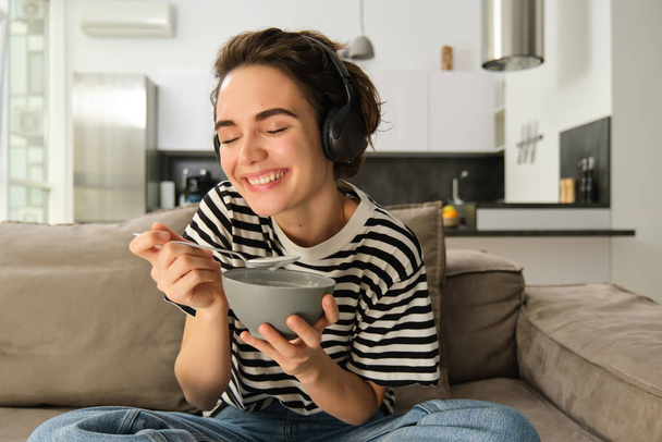 Primer plano de la hermosa mujer feliz, riendo y sonriendo, viendo la televisión en auriculares inalámbricos, sosteniendo tazón de cereales, desayunando en la sala de estar en el sofá. - Foto, Imagen