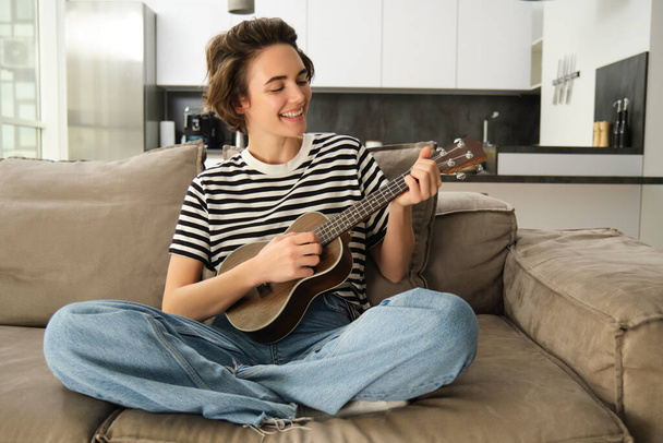 Koncepcja stylu życia i hobby. Młoda uśmiechnięta kobieta na kanapie, grająca na ukulele, śpiewająca i ucząca się wzorów do ulubionej piosenki. - Zdjęcie, obraz