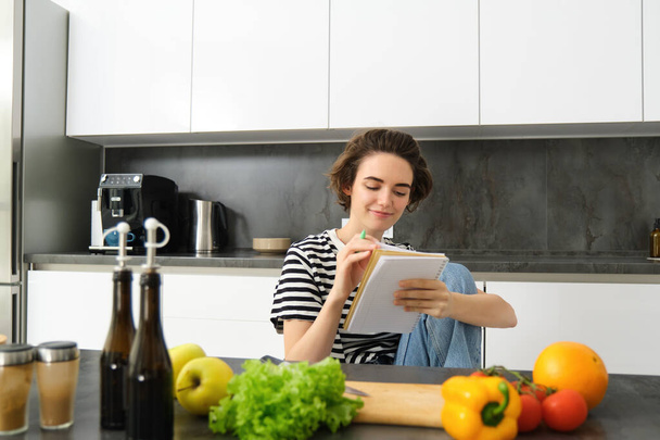 Portret młodej uśmiechniętej kobiety w kuchni, trzymając notatnik, robiąc notatki na przepis, pisząc listę zakupów, gotując sałatkę, siedząc przy warzywach i siekając deskę. - Zdjęcie, obraz