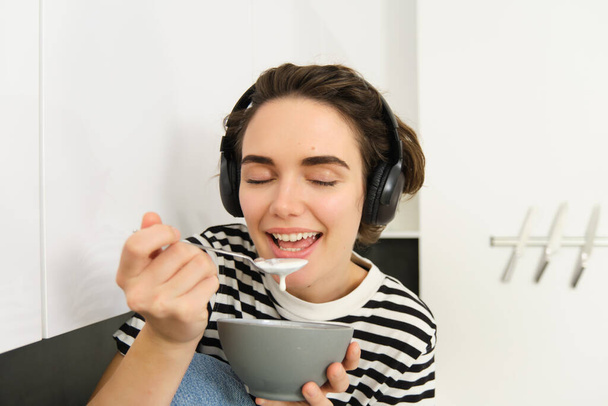 Close up portrait of young woman enjoys eating her breakfast, having cereals with milk, holding spoon and bowl, sits on kitchen counter with headphones, listens to music. - Photo, Image