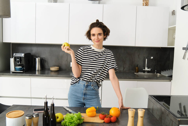 Portret pięknej, pewnej siebie uśmiechniętej kobiety, pozującej przy blacie kuchennym z warzywami i deską do krojenia, trzymającej jabłko, gotującej posiłek, przygotowującej sałatkę. - Zdjęcie, obraz