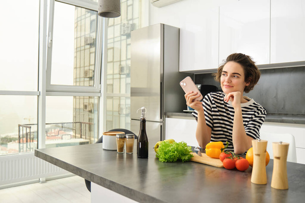 Portret van een jonge vrouw in de keuken met groenten en snijplank, smartphone vasthouden, gezonde dieetrecepten zoeken in de browser van de mobiele telefoon, thuis koken. - Foto, afbeelding