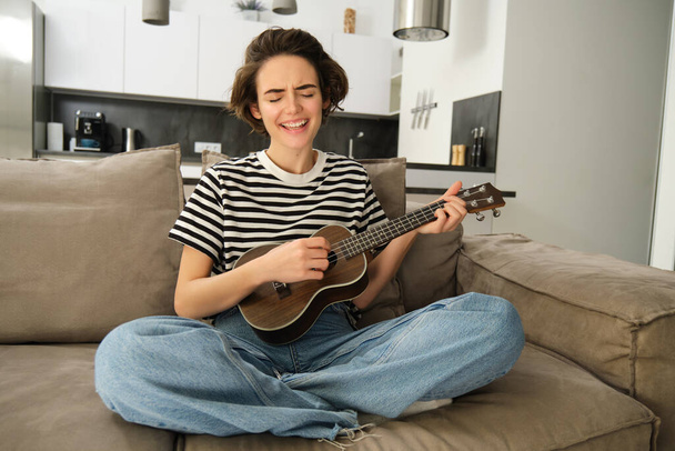 Portré aranyos fiatal nő, lány ukulele, játszik és énekel kedvenc dal, miközben ül a nappaliban kanapén, izgalmas húrok. - Fotó, kép