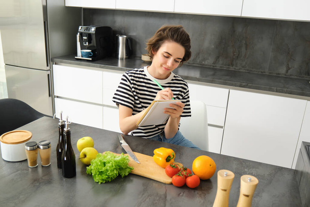 Portré nő írja le a receptjét, jegyzetel jegyzetfüzetbe főzés közben, ül a konyhában vágódeszkával és zöldségekkel, ételt készít. - Fotó, kép