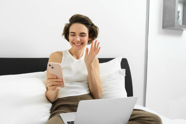 Obrázek krásné mladé ženy relaxující doma, sedí na posteli s notebookem a smartphonem, připojí se k video chatu, volá přítele, mává rukou na obrazovce mobilního telefonu a usmívá. - Fotografie, Obrázek