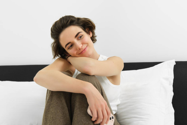 Jovem mulher macia em seu pijama acolhedor, sentada na cama no quarto, sorrindo com um sorriso sensual. - Foto, Imagem