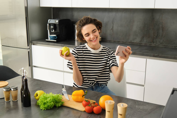 Jovencita sonriente, bloguera de comida grabando un video en un smartphone con comida, cocinando y haciendo selfie, tomando fotos durante la preparación de la comida, sentada cerca de verduras y sosteniendo una manzana. - Foto, imagen