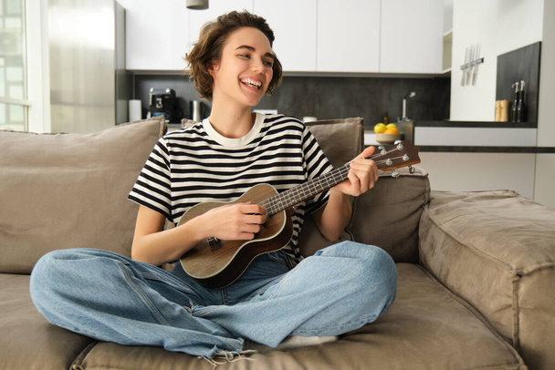 Wesoła młoda kobieta z ukulele, gra na instrumencie muzycznym, trzyma małą gitarę i śpiewa, siedzi na kanapie na skrzyżowanych nogach, odpoczywa w salonie. - Zdjęcie, obraz