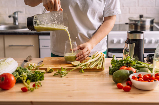 Dame in de keuken giet een smoothie in een glas op een houten tafel waarop verse groenten staan. - Foto, afbeelding