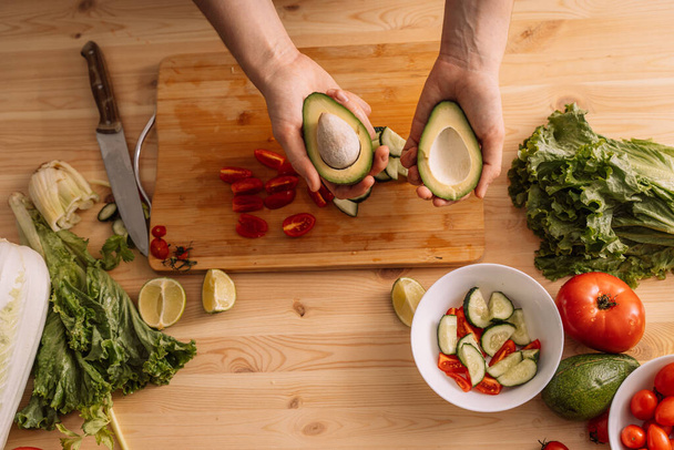 Lady halbiert eine Avocado auf einem Holztisch, auf dem frisches Gemüse liegt. - Foto, Bild