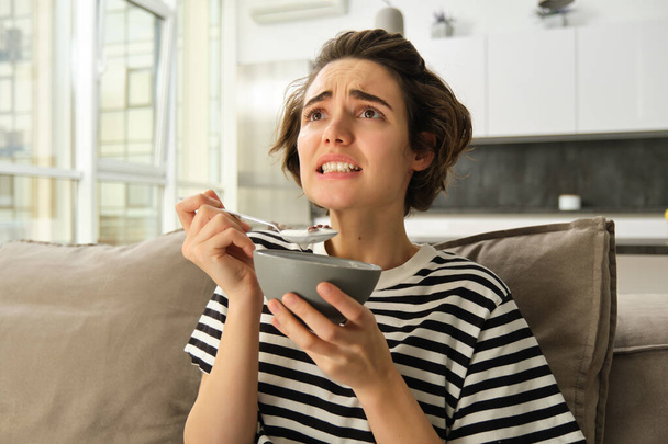 Image of woman eating breakfast in front of tv, looking at screen with worried face, holding bowl of cereals with milk and a spoon, spending time in living room. - Photo, Image