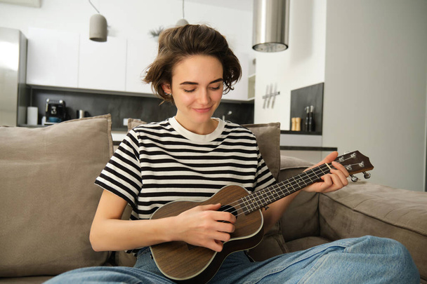 Portret van een jonge moderne vrouw, student die thuis ukelele speelt, met kleine gitaar zit, zingt en zich gelukkig voelt, zittend op de bank. Lifestyle en muziek concept - Foto, afbeelding