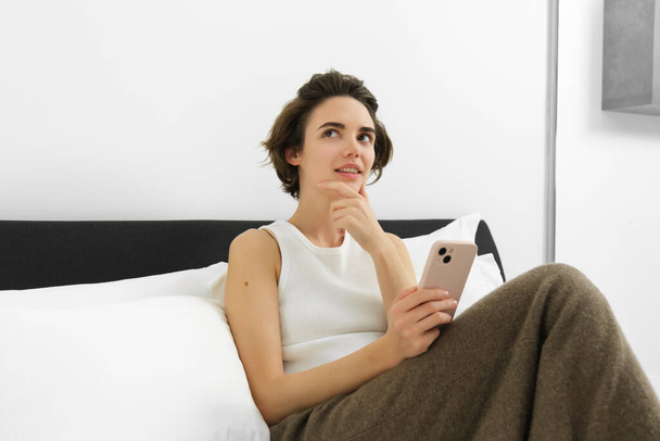 スマートフォン付きの美しい女性の肖像画は,ベッドに座っている間,携帯電話アプリでオンラインショッピングを決定し,考えの顔を作ります. - 写真・画像