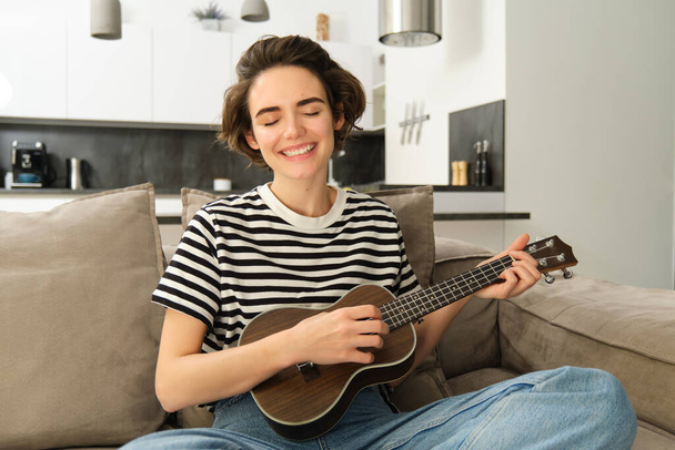 Young happy woman sitting on sofa and playing ukulele, singing and enjoying learning new musical instrument. - Photo, Image