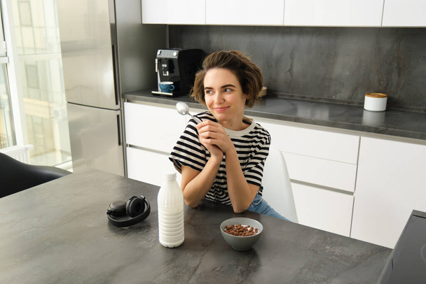 Portrait of smiling young candid woman, student eating morning cereals, having her breakfast, listening music in wireless headphones, sitting in the kitchen. - Photo, Image