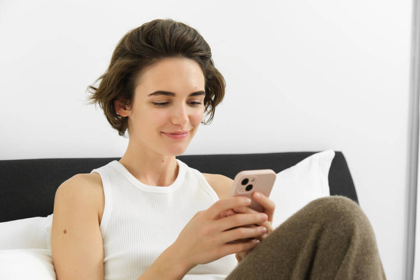 Ritratto di giovane bella donna bruna, seduta sul letto con smartphone, usando l'app per cellulare, inviando messaggi e sorridendo, leggendo sull'applicazione telefonica in camera da letto. - Foto, immagini