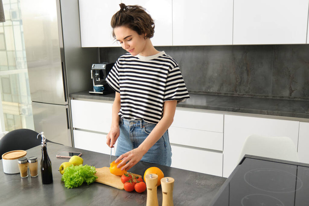 Πορτρέτο της σύγχρονης νεαρής γυναίκας που στέκεται στην κουζίνα με λαχανικά, κόβοντας τους στον πάγκο, κρατώντας μαχαίρι, φαγητό μαγειρικής, χορτοφαγικό φαγητό, έννοια του υγιεινού τρόπου ζωής μια διατροφή. - Φωτογραφία, εικόνα