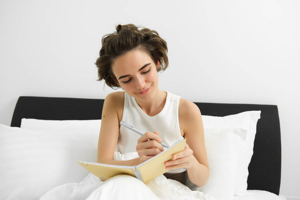 Kuva tietoisesta nuoresta naisesta, joka istuu sängyssä, herää ja kirjoittaa päiväkirjaan, tekee muistiinpanoja, jakaa ajatuksiaan päiväkirjaan. - Valokuva, kuva