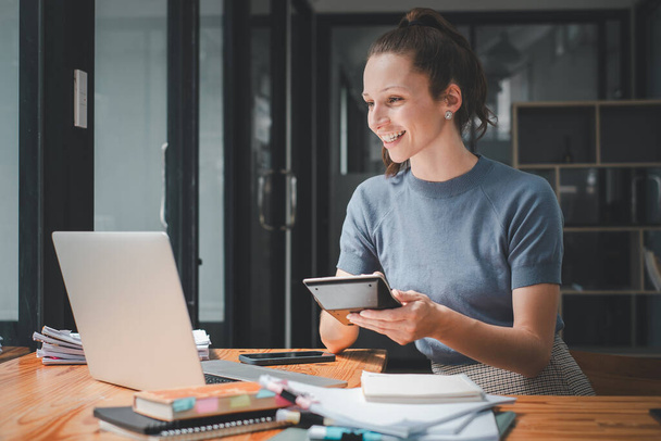 Lächelnde junge Frau, die mit Taschenrechner und Laptop Finanzbericht berechnet, im Büro mit Laptop auf dem Tisch arbeitet.  - Foto, Bild