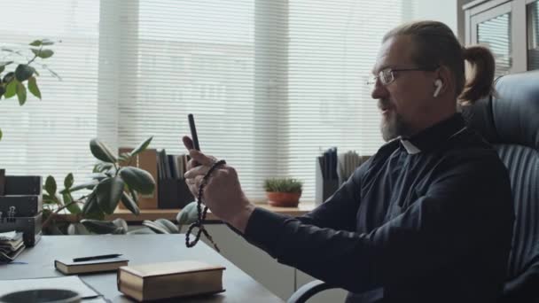 Zijaanzicht van volwassen katholieke priester met paardenstaart kapsel dragen oordopjes zitten aan houten bureau in zijn kantoor praten op video oproep op smartphone - Video