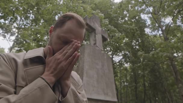 Foto de ángulo bajo del viudo con las manos rezando de luto mientras está sentado bajo el monumento de la cruz gris de hormigón en el cementerio - Metraje, vídeo