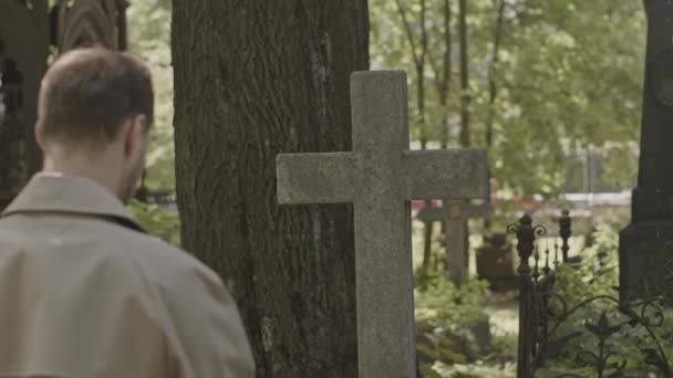 Powrót widok średni strzał mężczyzny w płaszczu wielbłąda stojącego przy krzyżu nagrobek na cichym cmentarzu w dzień - Materiał filmowy, wideo