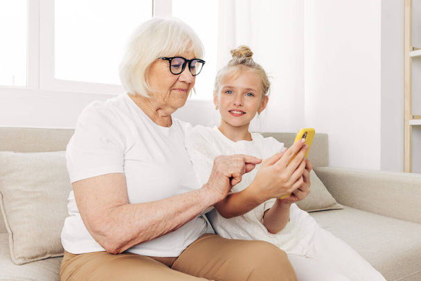 Grand-mère téléphone familial à l'intérieur enfant deux souriant vidéo selfie éducation t-shirt petite-fille collage photographie gens étreignant canapé espace copie ensemble appel blanc - Photo, image