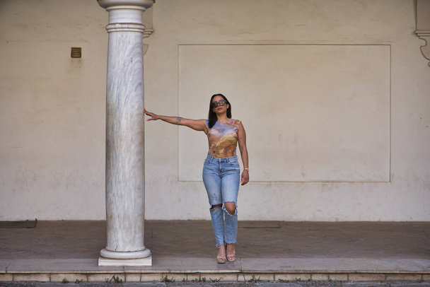 Young, beautiful, brunette and South American woman with a printed T-shirt and torn jeans, posing next to a marble column. Concept beauty, fashion, trend. - Photo, Image