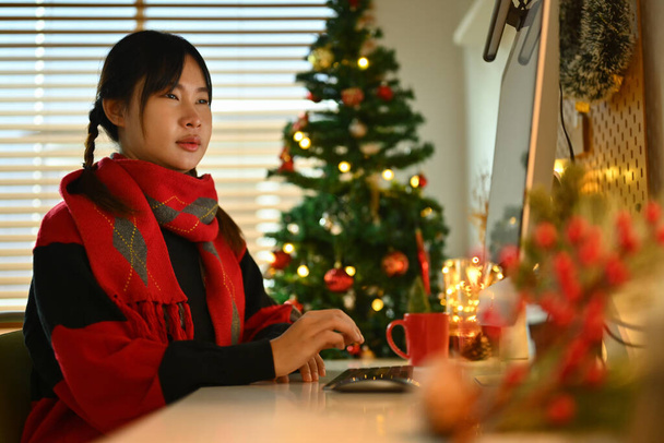 Junge Frau im Pullover blickt auf Computerbildschirm, während sie in dekoriertem Wohnzimmer für Weihnachten arbeitet. - Foto, Bild
