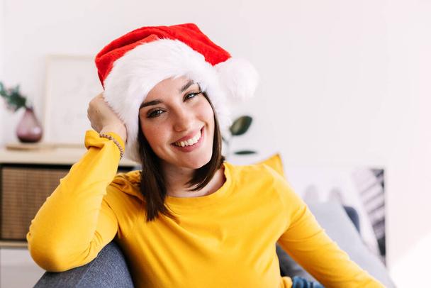 Дружній портрет молодої тисячолітньої жінки в капелюсі Діда Мороза, яка посміхається на камеру, розслабляючись на дивані на різдвяній відпустці вдома - Фото, зображення