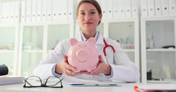 Γιατρός θεραπευτής κρατά κουμπαρά στην κλινική. Ιατρικές δαπάνες και ασφάλιση υγείας και εξοικονόμηση χρημάτων και κόστους - Πλάνα, βίντεο