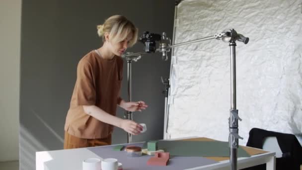 Medium shot van jonge blanke vrouwelijke fotografe die in de studio werkt, flatlay fotografeert met camera op horizontaal rek en geometrische vormen op tafel aanpast - Video