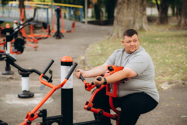 un uomo grasso beve e fa sport nel parco su simulatori - Foto, immagini