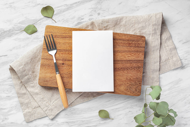 Деревянная доска и макет карточки меню с вилкой и льняной салфеткой на мраморном кухонном столе с зелеными листьями, вид сверху - Фото, изображение