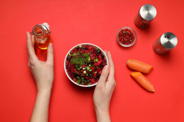 Салат винегрет в миске, морковь, специи и руки на красном фоне, вид сверху - Фото, изображение