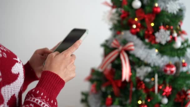 Nuori nainen, joka on upotettu puhelimeensa, tekstaa iloisesti joulun lomakauden aikana, lisäämällä juhlavan kosketuksen hänen elämäntapaansa männyn taustalla.. - Materiaali, video